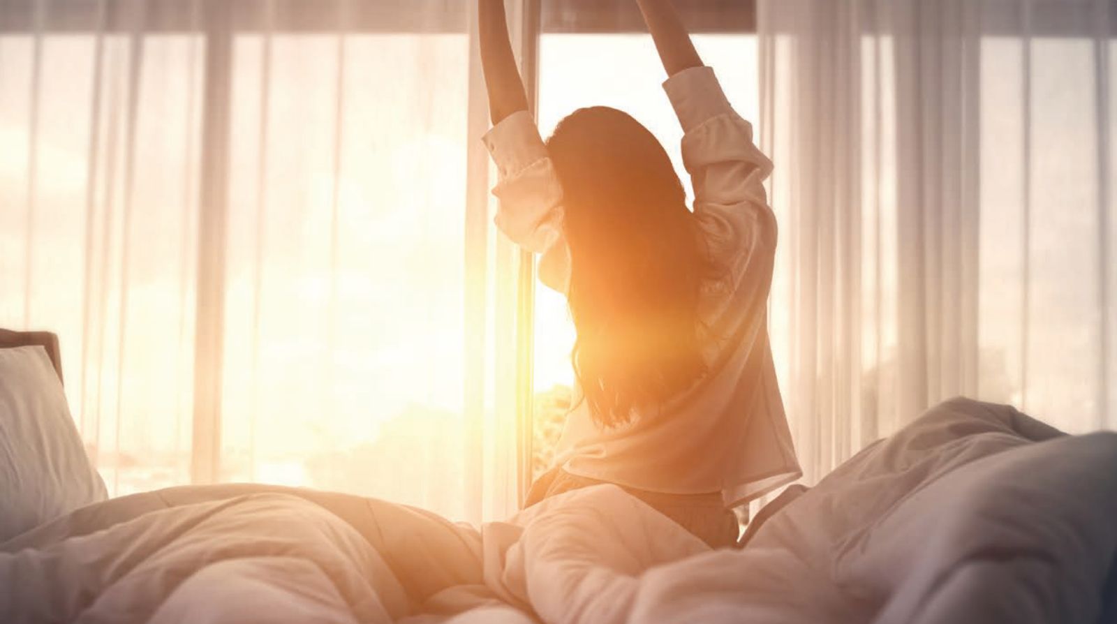 Frau steht aus dem Bett auf während Sonne durch das Fenster strahlt